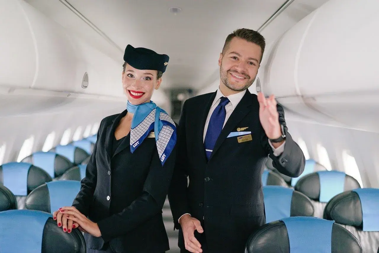 Uśmiechnięta stewardessa i steward w mundurach LOT stoją w samolocie, na tle rzędów siedzeń.