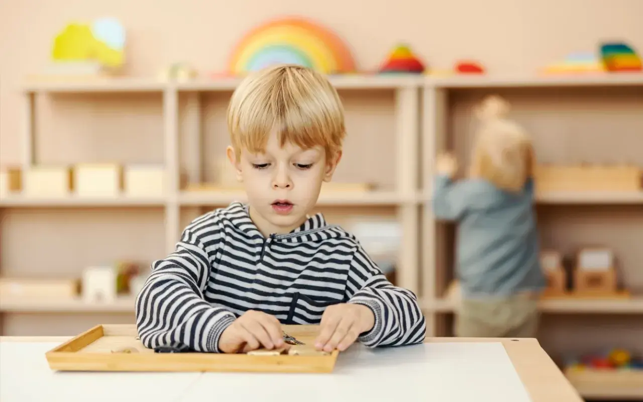 Chłopiec w bluzie w paski skupiony na zadaniu z materiałami Montessori. W tle inne dziecko przy półce.