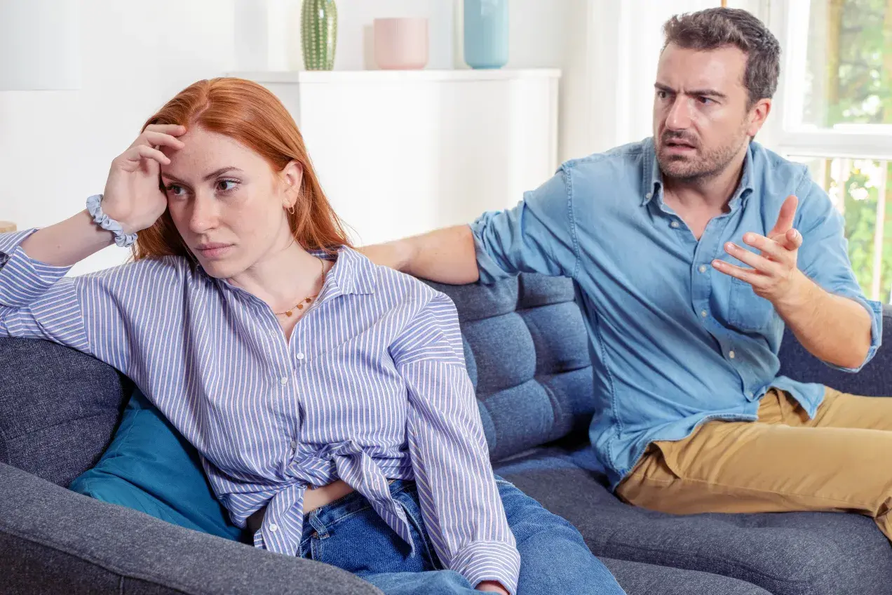 Para siedząca na kanapie, mężczyzna mówi z wyrzutem, kobieta odwrócona, zmartwiona.