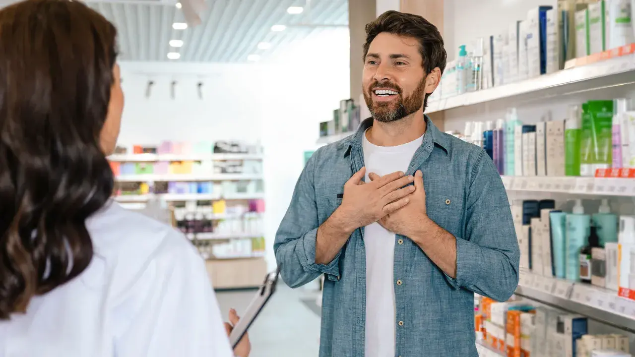 Mężczyzna z dłońmi na piersi rozmawia z farmaceutką, zastanawiając się, ile zarabia farmaceuta.