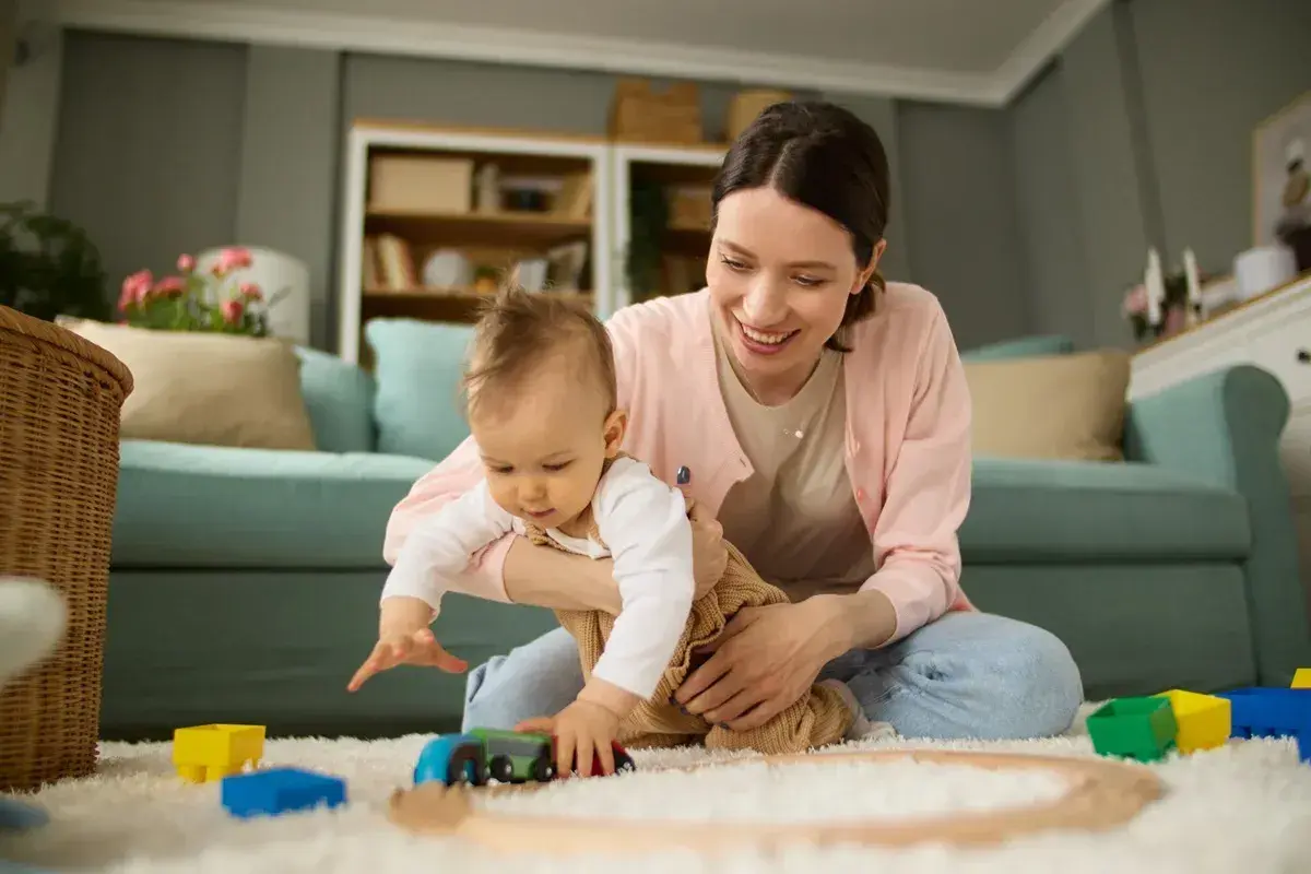 Uśmiechnięta mama bawi się z dzieckiem klockami i pociągiem na dywanie w salonie.