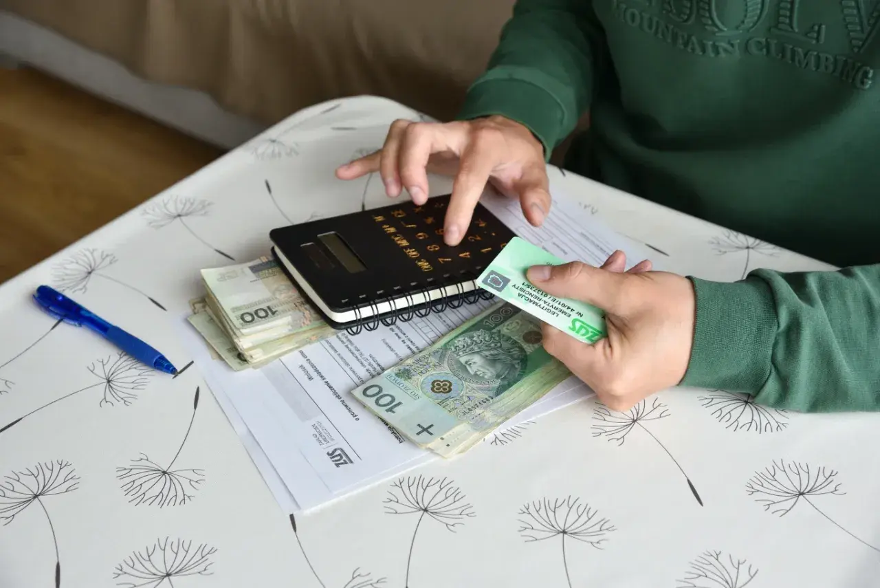 Dłonie trzymające pieniądze, kalkulator i dokumenty ZUS na stoliku.