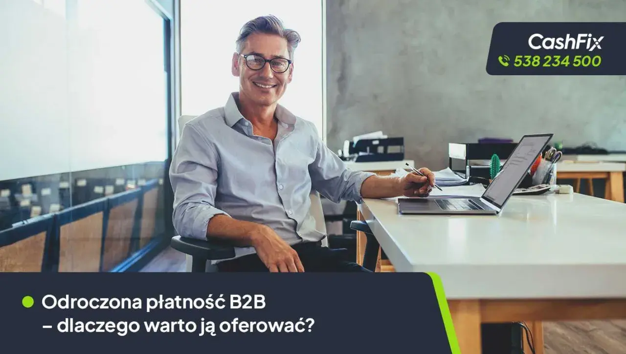 Uśmiechnięty mężczyzna w okularach siedzi przy biurku z laptopem, pisząc coś na papierze.