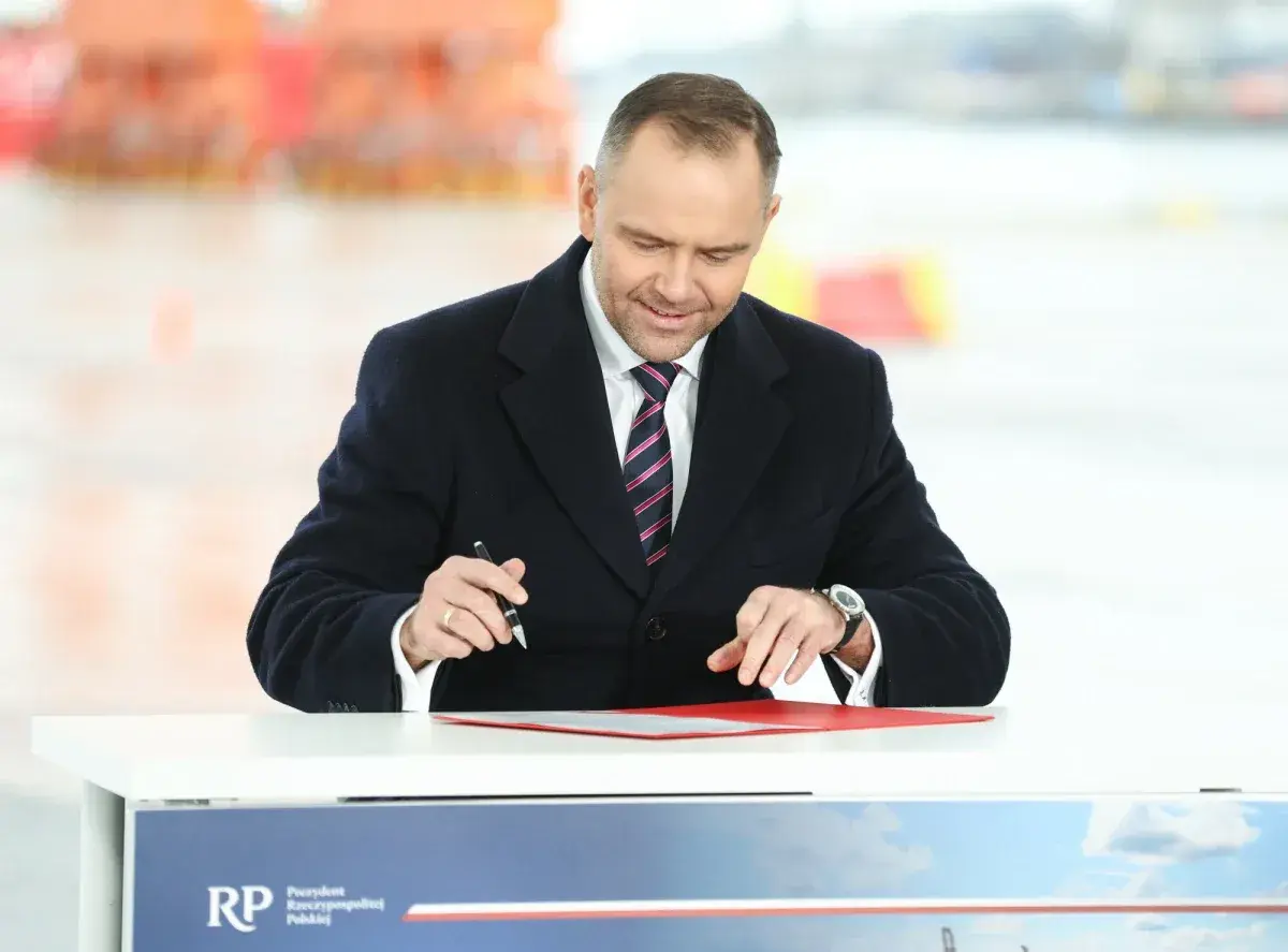 Mężczyzna w garniturze i płaszczu podpisuje dokumenty przy stole.