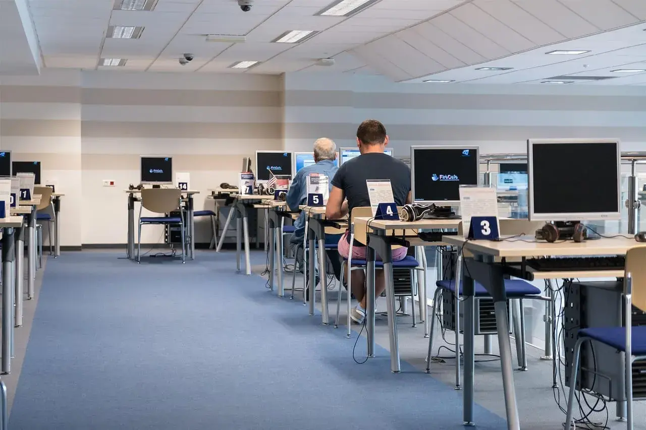 Rząd stanowisk komputerowych z osobami pracującymi, niebieska wykładzina.