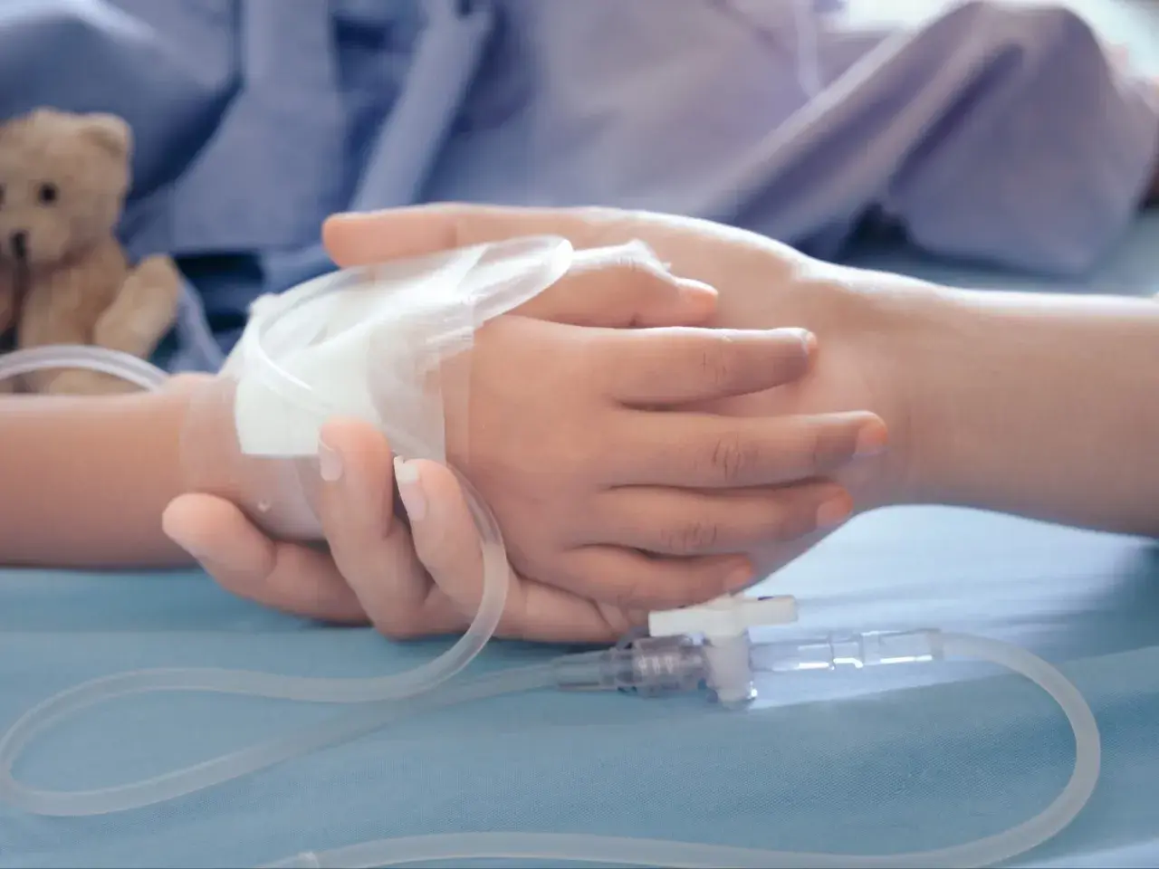 Dłoń dorosłego obejmuje dłoń dziecka z wenflonem. W tle pluszowy miś. Obraz symbolizuje wsparcie i troskę, jakiej wymaga zasilek pielegnacyjny.