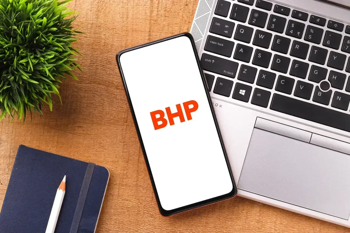 Telefon z napisem BHP na ekranie, obok laptopa, notatnika i rośliny.