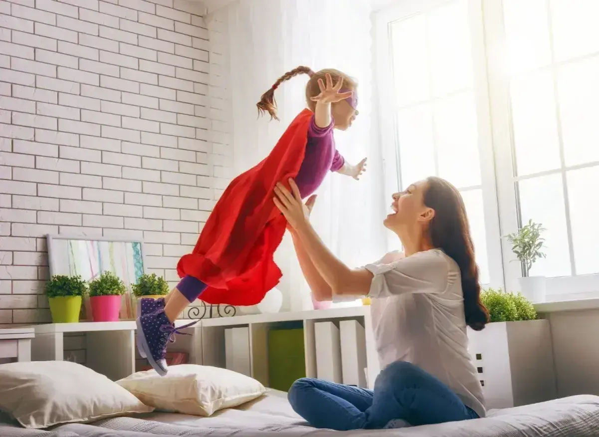Radosna dziewczynka w stroju superbohaterki lata w górę, złapana przez uśmiechniętą mamę. To wspaniałe, babciowe chwile pełne zabawy.