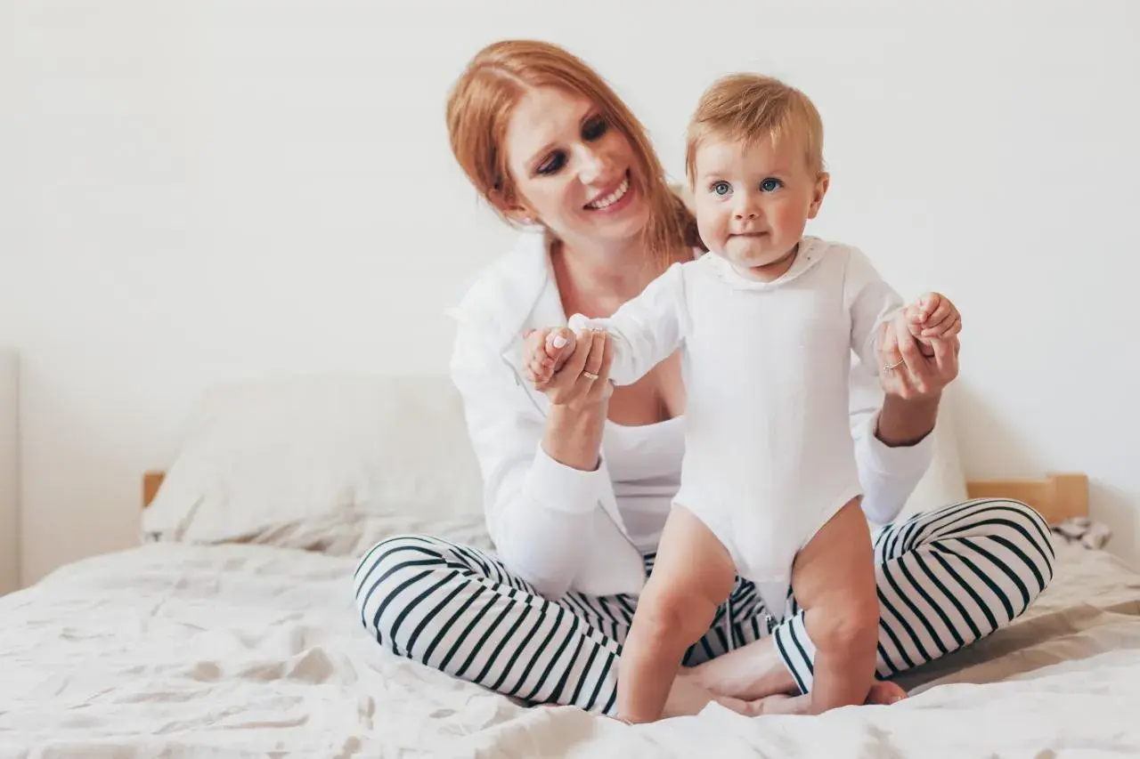 Uśmiechnięta mama z rudymi włosami pomaga 8-miesięcznemu dziecku w białym body wstać na łóżku.
