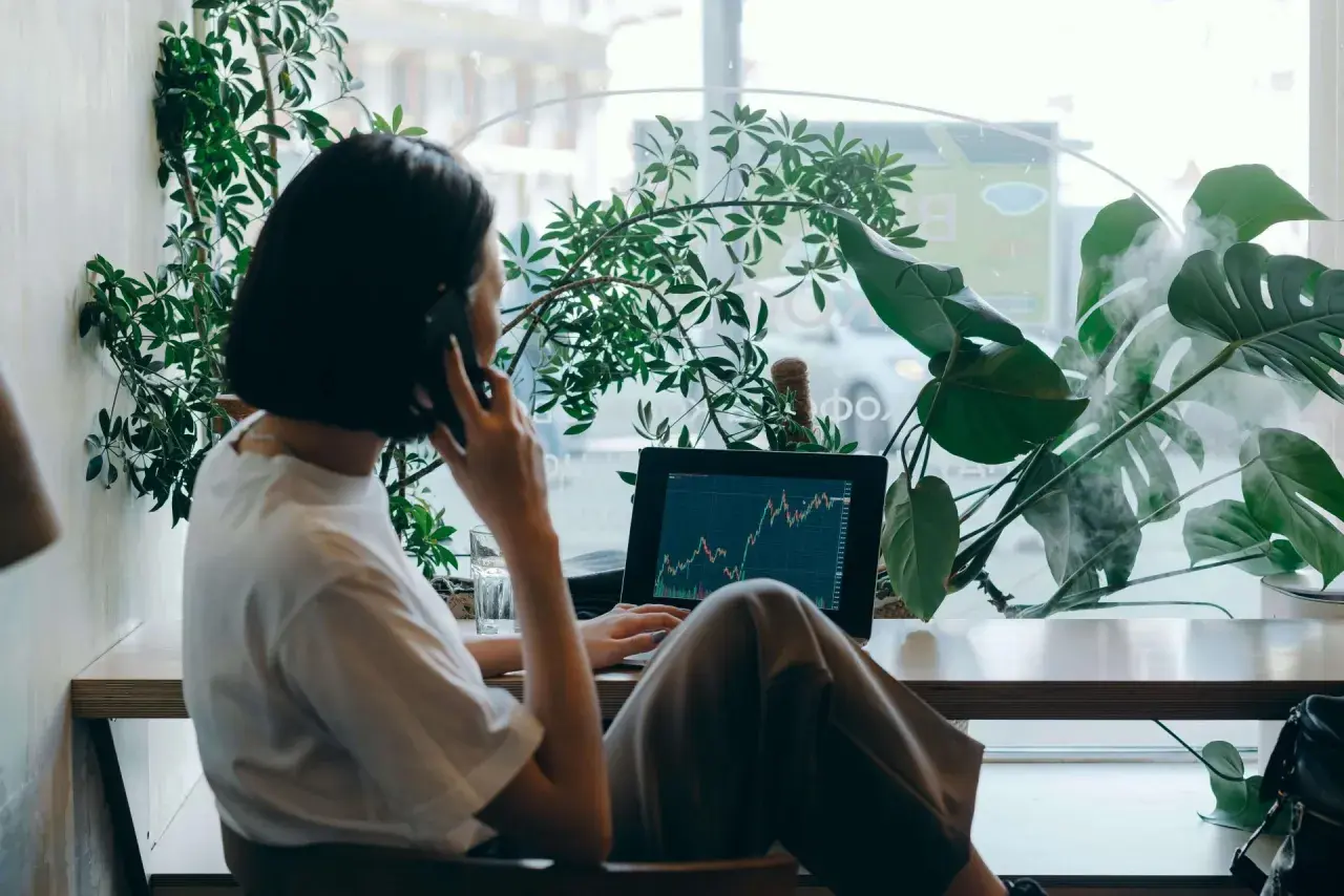 Kobieta rozmawia przez telefon przy laptopie z wykresem giełdowym, otoczona roślinami.