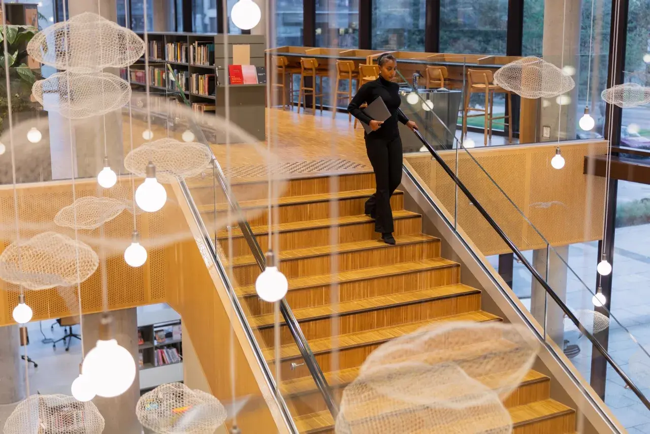 Kobieta z laptopem schodzi po drewnianych schodach w nowoczesnej bibliotece z ozdobnymi chmurkami i lampami.