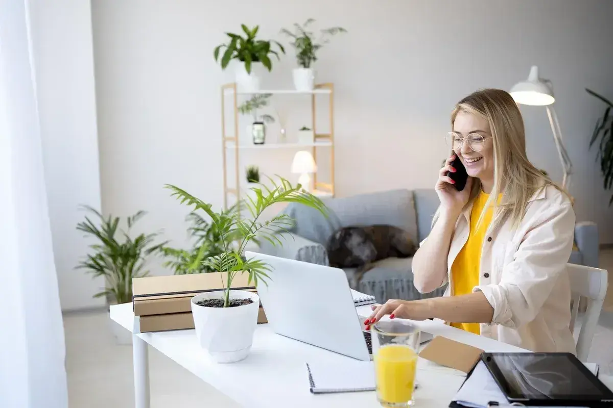 Uśmiechnięta kobieta pracuje przy laptopie i rozmawia przez telefon w domowym biurze.