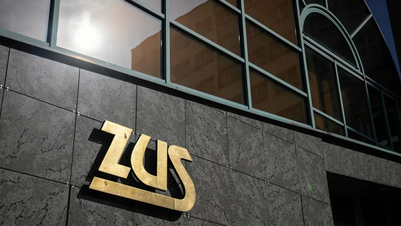 Złote logo ZUS na ciemnej, kamiennej elewacji budynku, nad nim okna z odbiciem.