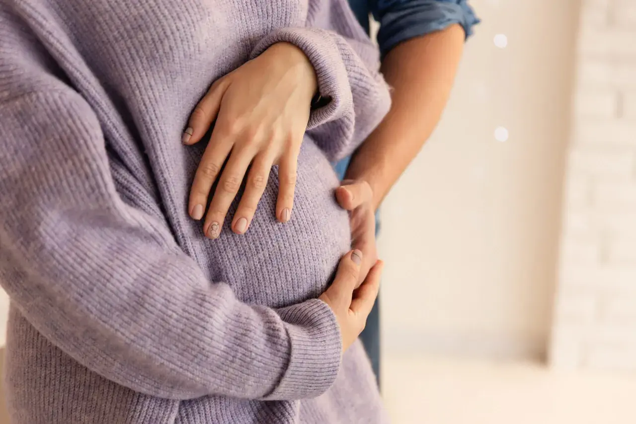 Dłoń kobiety w fioletowym swetrze obejmuje ciążowy brzuch, obok dłoń partnera.