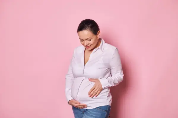 Uśmiechnięta kobieta w ciąży po 40. delikatnie dotyka brzucha na różowym tle.